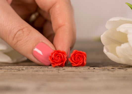 Red rose-shaped earrings held between fingers with a white flower in the background.