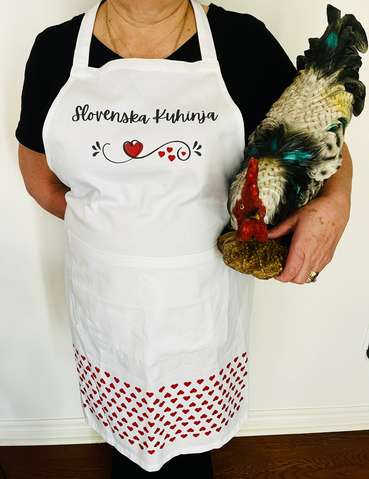 A person wearing a white cotton apron with red patterns and the text 'Slovenska Kuhinja' printed on it, holding a chicken figurine.